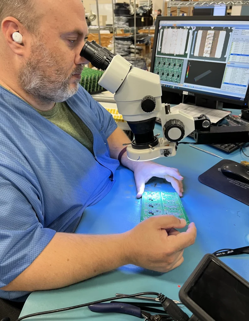 Technician examining a populated circuit board under a microscope at an inspection workstation